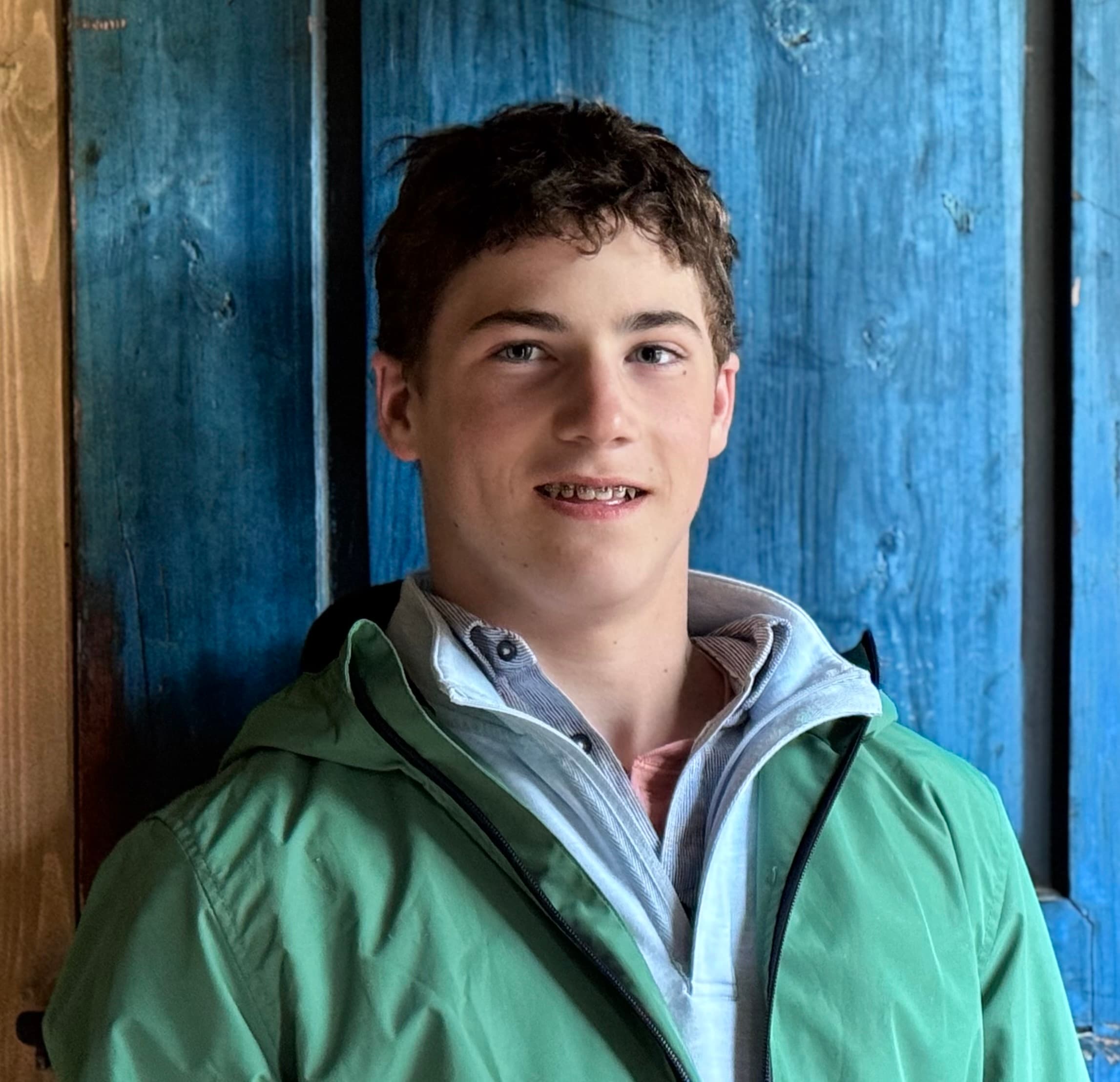 Smiling teenage boy with curly hair and braces wearing a green jacket against blue wood.
